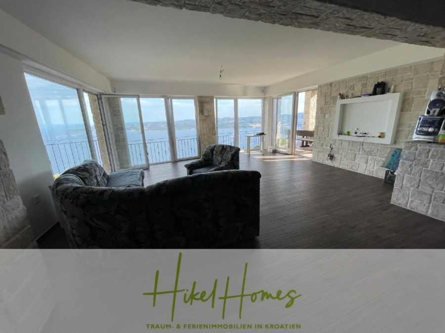 Meerblick aus perfekter Höhenlage mit 3 Schlafzimmern - Geräumiges, modernes Wohnzimmer mit Steinwänden, großen Fenstern mit Panoramablick aufs Meer, dunklen Holzböden und zwei gemusterten Sofas. Der Text "Hikel Homes - Traum- & Ferienimmobilien in Kroatien" erscheint am unteren Rand. (46057)