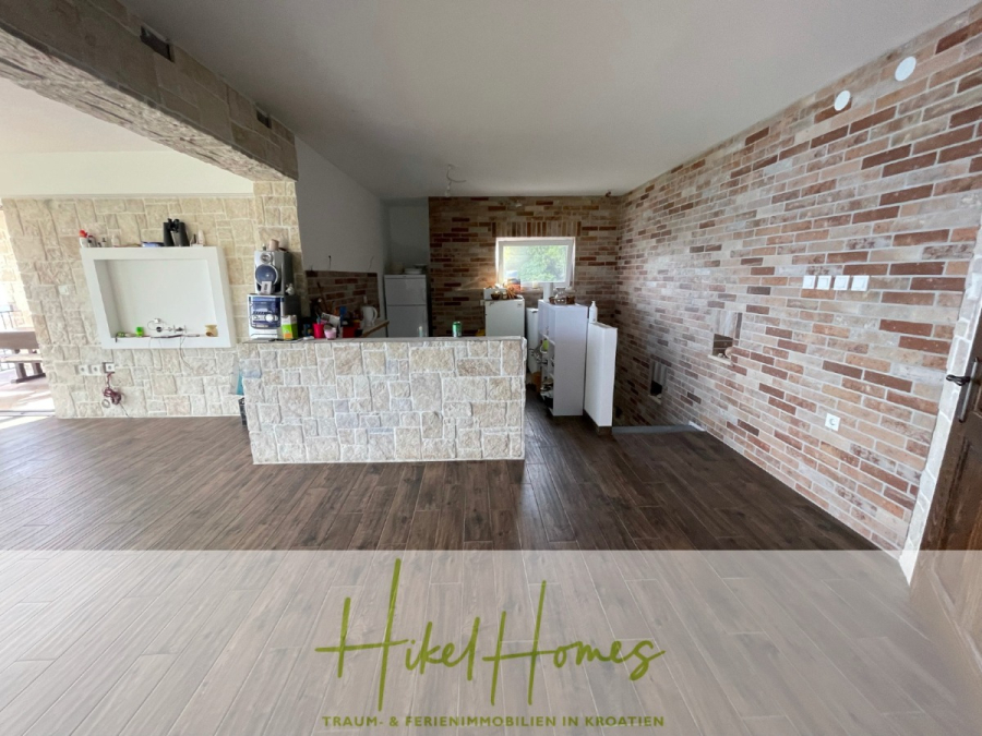 Meerblick aus perfekter Höhenlage mit 3 Schlafzimmern - Eine geräumige Küche mit Stein- und Ziegelwänden, Holzfußboden, weißen Schränken, einem kleinen Kühlschrank und verschiedenen Küchengeräten auf dem Tresen. Große Fenster lassen natürliches Licht herein. Der Schriftzug "Hikel Homes" erscheint am unteren Rand. (46065)