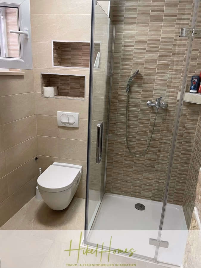Meerblick aus perfekter Höhenlage mit 3 Schlafzimmern - Modernes Badezimmer mit verglaster Dusche, beigefarbenen Fliesen, Wand-WC und Einbauregalen. Über dem WC befindet sich ein kleines Fenster. Braun- und Beigetöne sorgen für eine warme Atmosphäre. (41466)