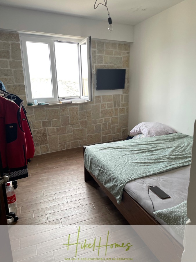 Meerblick aus perfekter Höhenlage mit 3 Schlafzimmern - Ein kleines Schlafzimmer mit einem Bett, grauem Bettzeug, einem an der Wand montierten Fernseher, einem Fenster, das Tageslicht hereinlässt, und einer Tapete mit Steinmustern. Ein Kleiderständer mit Jacken und ein Feuerlöscher stehen in der Ecke. Das "HikeHomes"-Logo befindet sich am Boden. (46061)