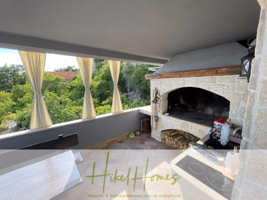 Meerblick aus perfekter Höhenlage mit 3 Schlafzimmern - Außenküche mit Steinofen, Brennholzstapel und Herd. Cremefarbene Vorhänge hängen von den offenen Wänden und bieten einen Blick auf üppige grüne Bäume. Ein Logo und eine Textüberlagerung am unteren Rand verweisen auf „HikeHomes“ und Immobilien in Kroatien. (41483)