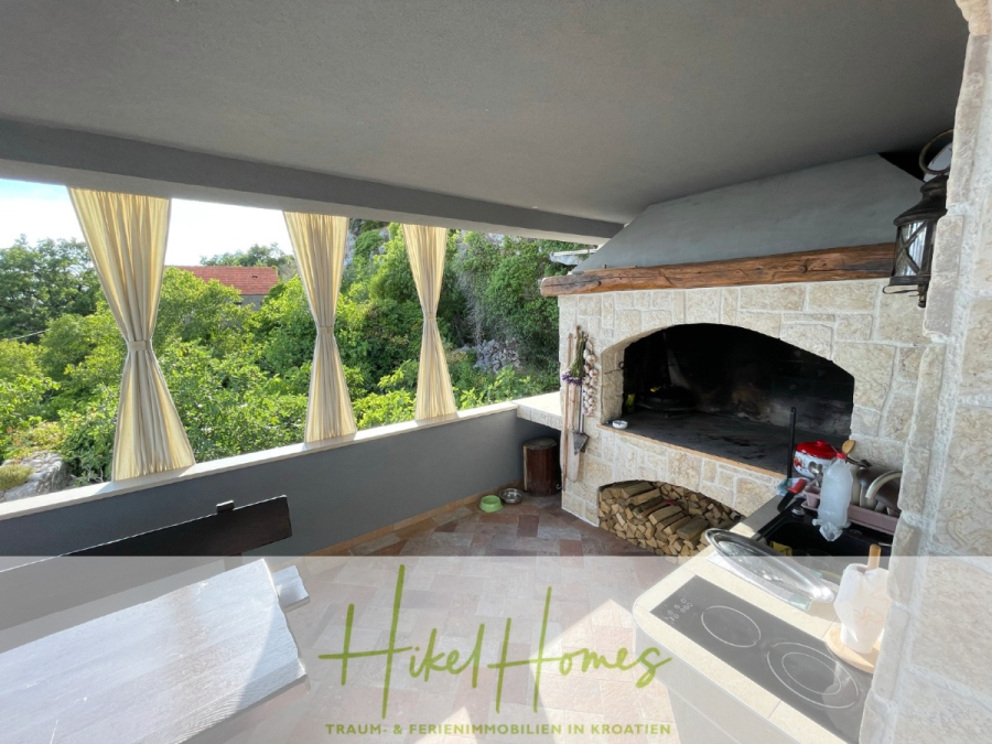 Meerblick aus perfekter Höhenlage mit 3 Schlafzimmern - Steinküche im Freien mit eingebautem Grill, Holzlager und Herd, umgeben von hellen Vorhängen und mit Blick ins Grüne. Ein weißer Tisch steht im Vordergrund. Das Textbanner "Hikel Homes" befindet sich am unteren Rand. (46082)