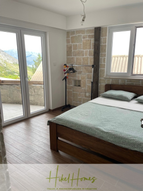 Ein helles Schlafzimmer mit einem Doppelbett, grüner Bettwäsche, einem kleinen Kleiderständer, gefliesten Wänden und großen Fenstern, die auf einen Balkon mit Bergblick führen. Der Boden ist aus Holz und der Raum wirkt gemütlich und luftig. - Meerblick aus perfekter Höhenlage mit 3 Schlafzimmern