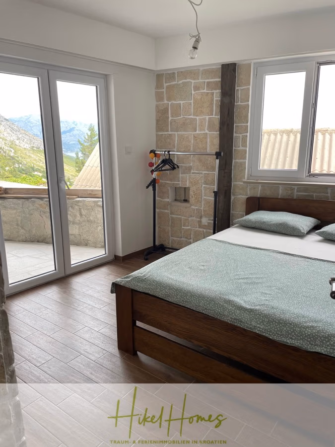 Meerblick aus perfekter Höhenlage mit 3 Schlafzimmern - Schlafzimmer mit Doppelbett, hellgrüner Bettwäsche und Kleiderständer. Große Glastüren und ein Fenster bieten vom gemauerten Balkon aus einen Blick auf die Berglandschaft. Der Boden ist gefliest, und an der Decke hängen Deckenleuchten. Das Logo lautet „HikelHomes“. (41464)
