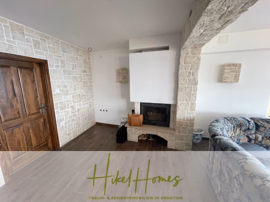 Meerblick aus perfekter Höhenlage mit 3 Schlafzimmern - Ein gemütliches Wohnzimmer mit Steinwänden, einem Kamin mit weißem Schornstein und umgebenden Steinelementen. Rechts steht ein Sofa mit Blumenmuster. Links eine Holztür. Fliesenboden ergänzt den rustikalen Stil. (41467)