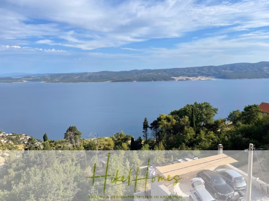 Meerblick aus perfekter Höhenlage mit 3 Schlafzimmern - Malerische Aussicht auf ein ruhiges Meer mit bergiger Küste unter leicht bewölktem Himmel. Im Vordergrund sind Grünflächen, einige Bäume und geparkte Autos zu sehen. Unten steht „Hikel Homes“. (41491)