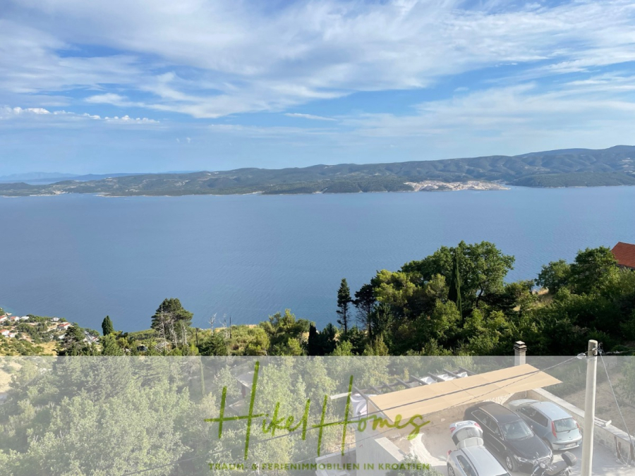 Meerblick aus perfekter Höhenlage mit 3 Schlafzimmern - Blick auf ein ruhiges blaues Meer mit Bergen in der Ferne, grünen Bäumen im Vordergrund und mehreren geparkten Autos. Der Himmel ist teilweise bewölkt, und der Text am unteren Rand lautet "Hike!Homes" mit einem Untertitel auf Deutsch. (46093)