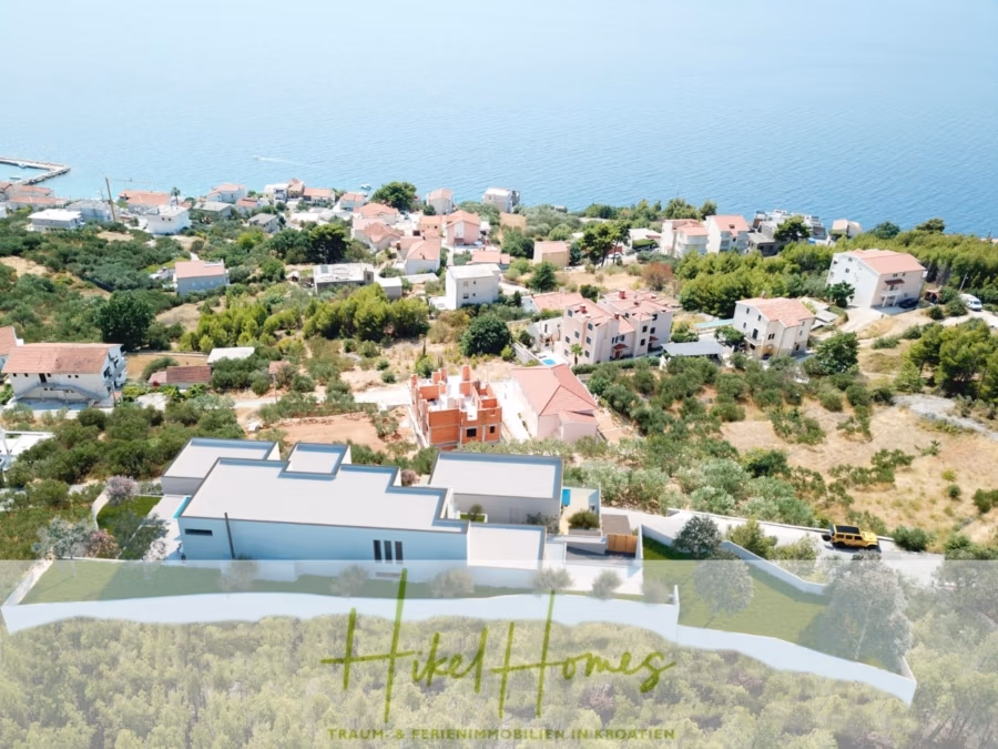 Extraklasse Meerblick Villa Three-Of-Three / Pool, 4 SZ, Lift, Konoba, Fitness, … - Luftaufnahme eines Küstendorfes mit rot gedeckten Häusern in der Nähe eines Gewässers. Im Vordergrund steht ein modernes graues Gebäude, umgeben von Grün. Das Wasser erstreckt sich bis zum Horizont. (42081)