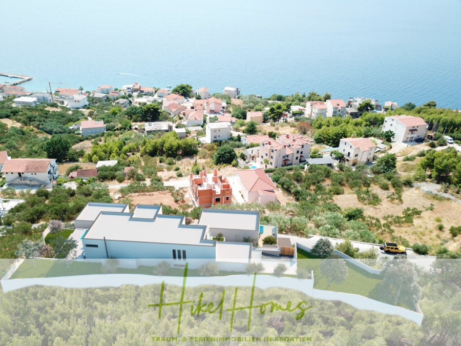 Extraklasse Meerblick Villa Three-Of-Three / Pool, 4 SZ, Lift, Konoba, Fitness, … - Luftaufnahme eines Küstendorfs mit Häusern inmitten von Grün in der Nähe des Meeres; im Vordergrund ein modernes Gebäude mit dem Text "Hikel Homes" und dem Slogan, der unten eingeblendet ist. (44095)