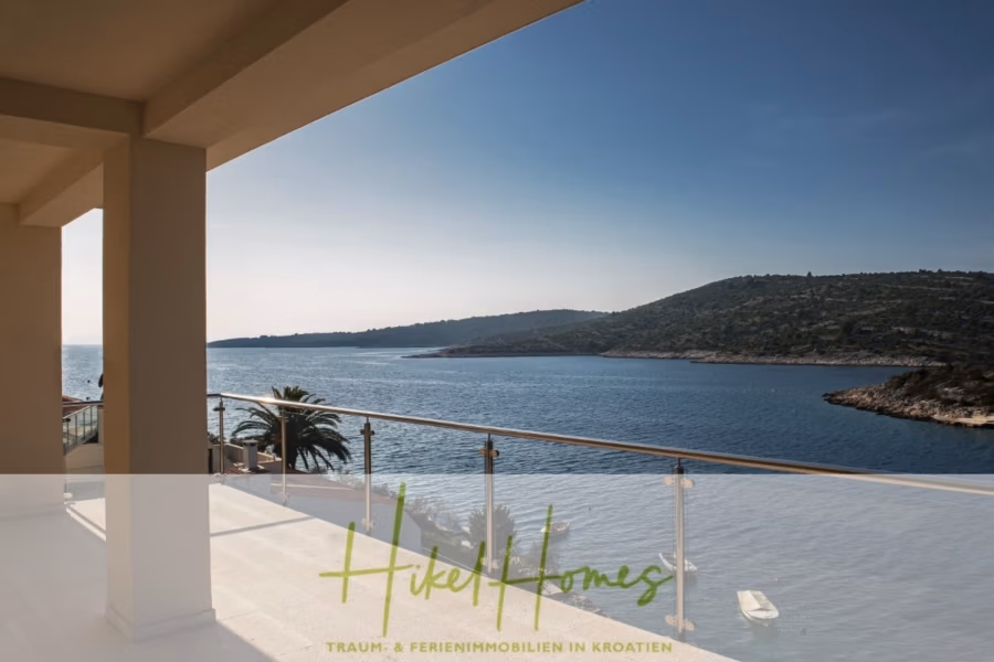 Villa Ana – 250m² WFL in 1. Reihe zum Meer / 4 SZ / Neubau / Sofort bezugsfertig - Blick vom Balkon auf eine ruhige Bucht mit klarem, blauem Wasser und weit entfernten, üppigen Hügeln unter klarem Himmel. Der Balkon hat ein Glasgeländer und in der Nähe steht eine Palme. Der Text unten lautet: „Hikel Homes – Traum- & Ferienimmobilien in Kroatien“. (41543)