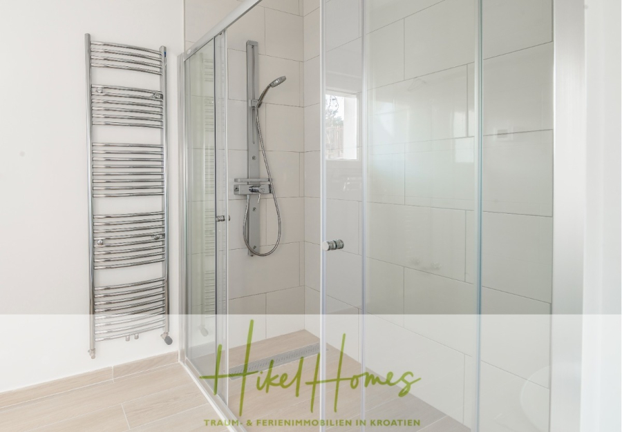 Villa Ana – 250m² WFL in 1. Reihe zum Meer / 4 SZ / Neubau / Sofort bezugsfertig - Modernes Badezimmer mit einer gläsernen Dusche, einem verchromten Handtuchheizkörper an der Wand, hellen Fliesen und einer Handbrause. Der Text am unteren Rand lautet "Hikel Homes Traum-& Ferienimmobilien in Kroatien. (46542)