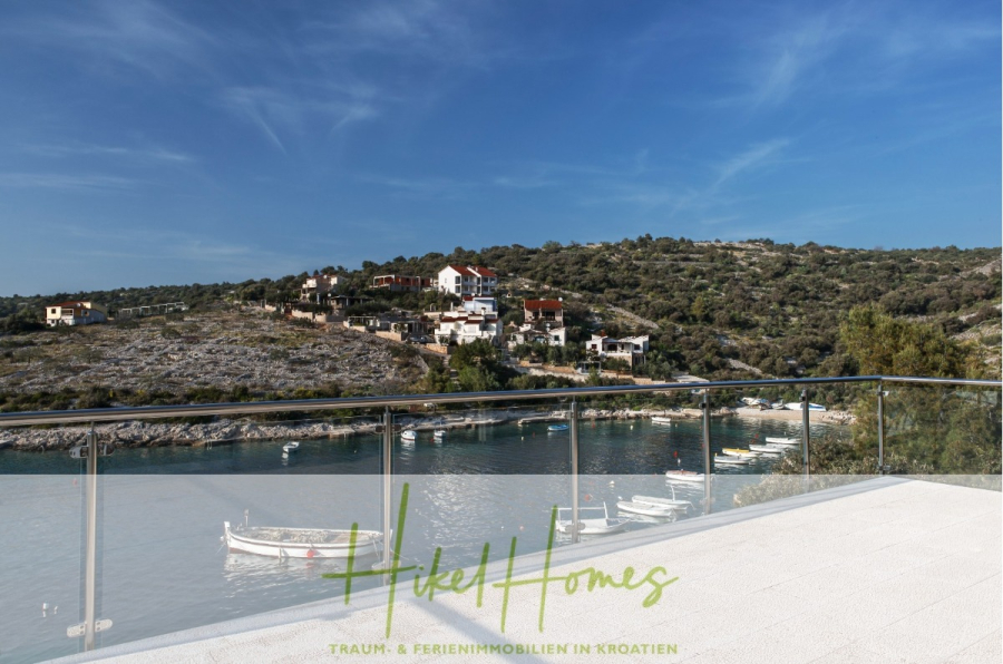 Villa Ana – 250m² WFL in 1. Reihe zum Meer / 4 SZ / Neubau / Sofort bezugsfertig - Blick von einer Terrasse mit Glasgeländer auf eine ruhige Bucht, kleine Boote auf dem Wasser und Häuser am Hang unter einem klaren blauen Himmel. Der Text "Hikel Homes" und "Traum- & Ferienimmobilien in Kroatien" befindet sich am unteren Rand. (46532)