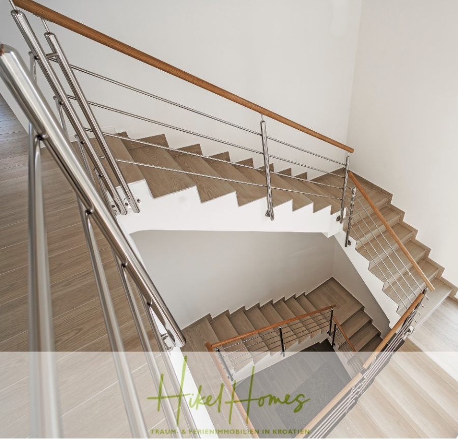 Villa Ana – 250m² WFL in 1. Reihe zum Meer / 4 SZ / Neubau / Sofort bezugsfertig - Moderne Innentreppe mit hellen Holzstufen, Metallgeländern und Holzhandläufen, von oben gesehen in einem sauberen, hellen Raum. Das Branding von Hikel Homes ist am unteren Rand des Bildes sichtbar. (46540)