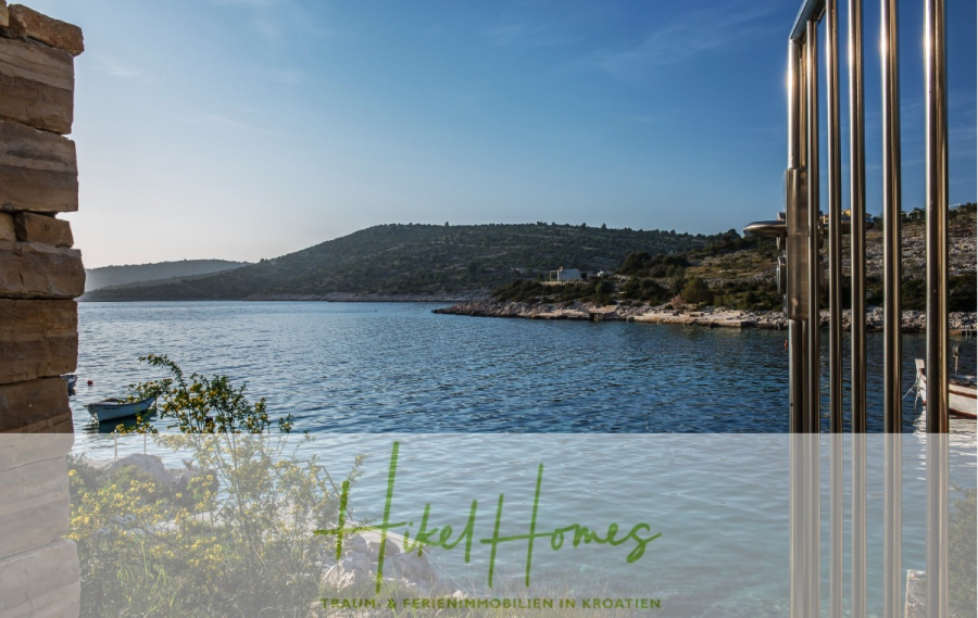 Villa Ana – 250m² WFL in 1. Reihe zum Meer / 4 SZ / Neubau / Sofort bezugsfertig - Blick von einer Steinterrasse mit Metallgeländer auf eine ruhige Bucht und entfernte Hügel unter einem klaren blauen Himmel. Ein kleines Boot schwimmt auf dem Wasser. Der Text lautet: "Heikel Homes - Traum & Ferienimmobilien in Kroatien.". (46536)