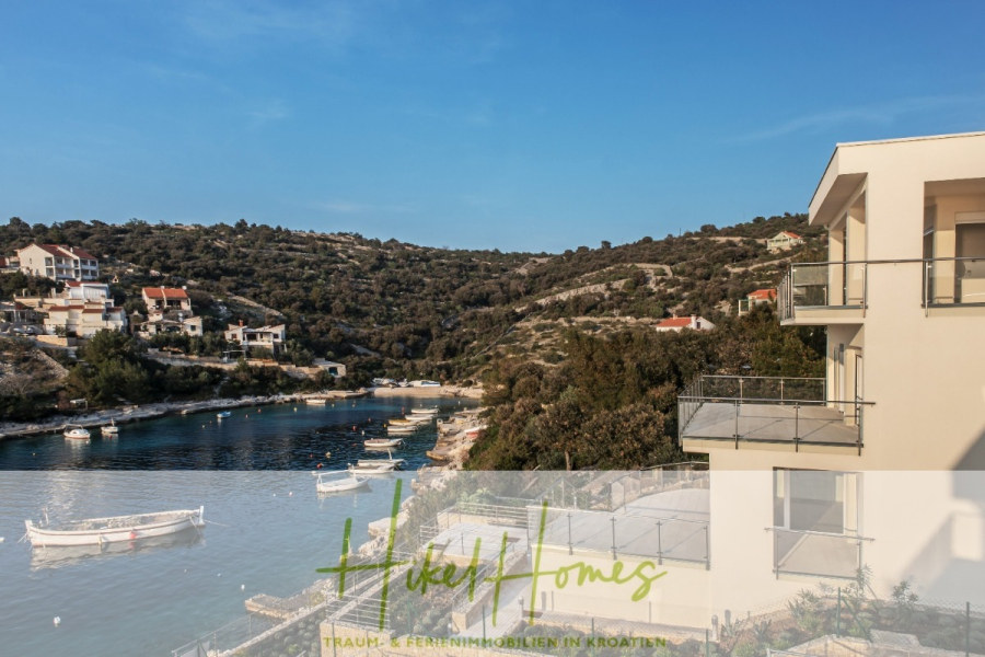 Villa Ana – 250m² WFL in 1. Reihe zum Meer / 4 SZ / Neubau / Sofort bezugsfertig - Blick auf eine friedliche Bucht mit kleinen Booten, die in Ufernähe vor Anker liegen, umgeben von grünen Hügeln und modernen Häusern. Im Vordergrund rechts ist ein beiges Gebäude mit Balkonen zu sehen. (46521)