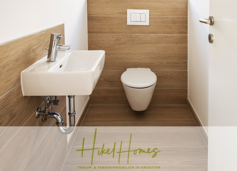 Villa Ana – 250m² WFL in 1. Reihe zum Meer / 4 SZ / Neubau / Sofort bezugsfertig - Ein kleines modernes Badezimmer mit Wänden im Holzstil, einem weißen Wandwaschbecken mit freiliegenden Rohrleitungen auf der linken Seite und einer weißen Wandtoilette. Auf der rechten Seite ist eine teilweise geöffnete Tür zu sehen. Das Logo "Hikel Homes" ist unten eingeblendet. (46541)