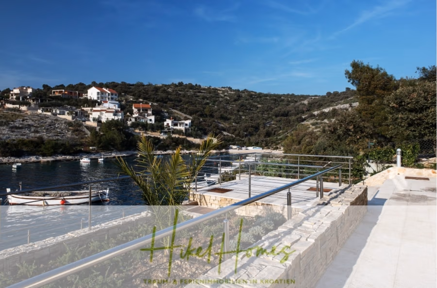 Villa Ana – 250m² WFL in 1. Reihe zum Meer / 4 SZ / Neubau / Sofort bezugsfertig - Malerischer Blick auf eine Küstenregion mit einer modernen Terrasse am Wasser und Blick auf eine ruhige Bucht. Boote liegen am felsigen Ufer vor Anker, im Hintergrund sind Häuser auf einem Hügel unter einem klaren blauen Himmel zu sehen. (41556)