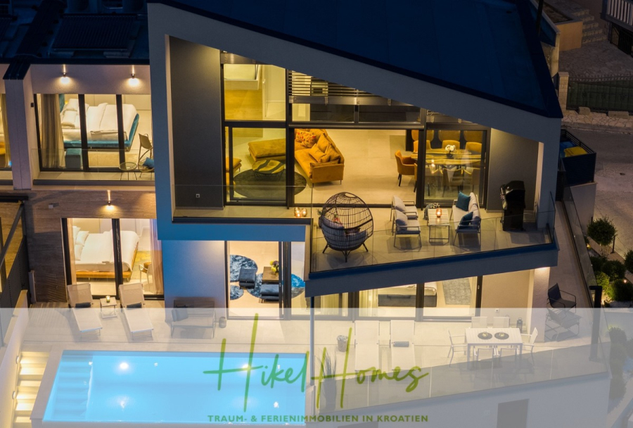 Direkt einziehen Meerblick Villa NATALIA mit Infinity Pool, 3 SZ, Sommerküche, … - Eine moderne, beleuchtete Villa bei Nacht mit großen Glasfenstern, sichtbaren Schlafzimmern, einem Wohnbereich und einem Swimmingpool. Das „Hike Homes“-Logo und der deutsche Text erscheinen am unteren Bildrand. (43871)