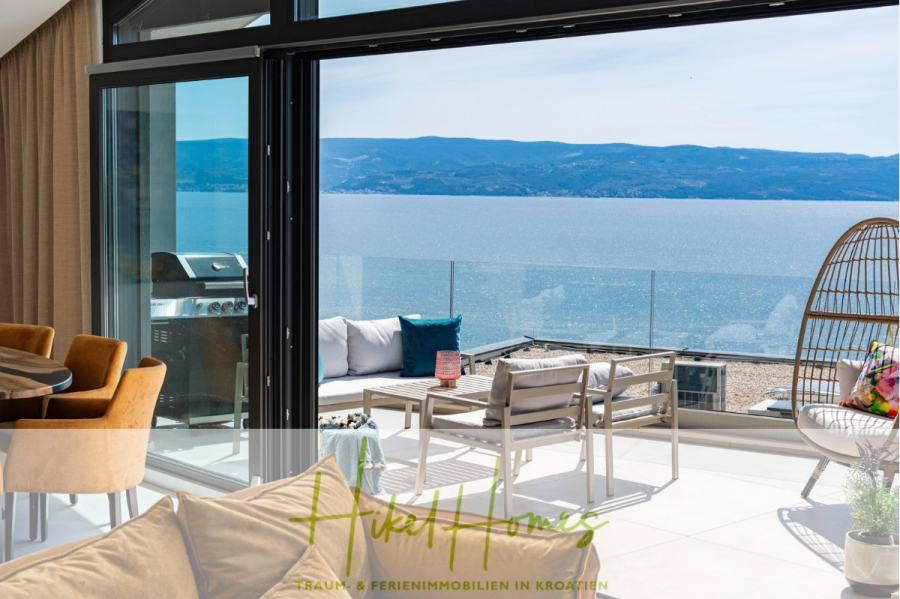 Direkt einziehen Meerblick Villa NATALIA mit Infinity Pool, 3 SZ, Sommerküche, … - Moderner, offener Wohnraum mit Glastüren, die auf eine Terrasse mit Blick auf das ruhige, blaue Meer und die Berge führen. Sitzgelegenheiten im Freien unter klarem Himmel. Unten steht „Akil Homes“. (43874)
