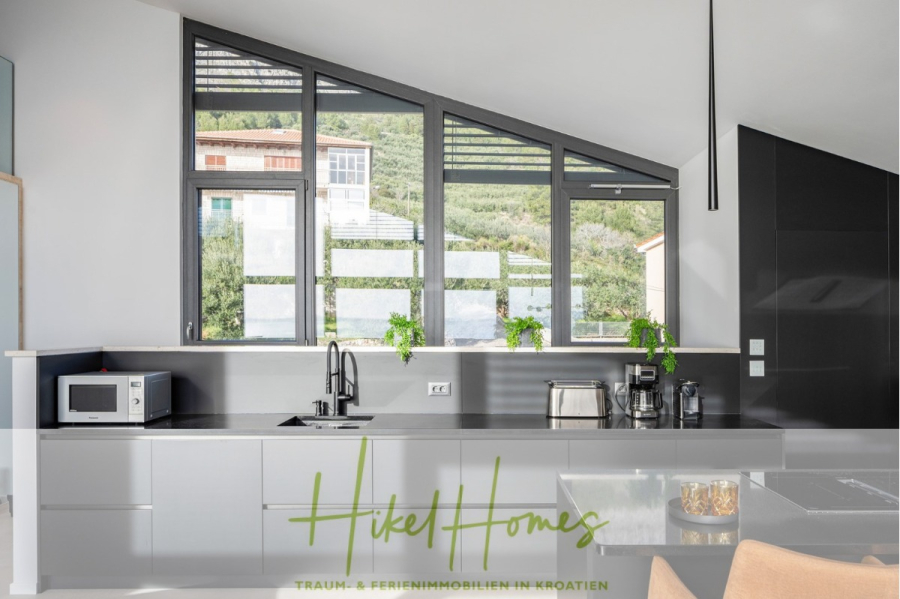 Direkt einziehen Meerblick Villa NATALIA mit Infinity Pool, 3 SZ, Sommerküche, … - Moderne Küche mit grauen Schränken, Einbaugeräten und großen Fenstern, die viel Tageslicht hereinlassen. Grüne Pflanzen stehen auf der Fensterbank, und draußen ist ein Hang zu sehen. Unten ist das Logo von „Hikel Homes“ zu sehen. (43869)