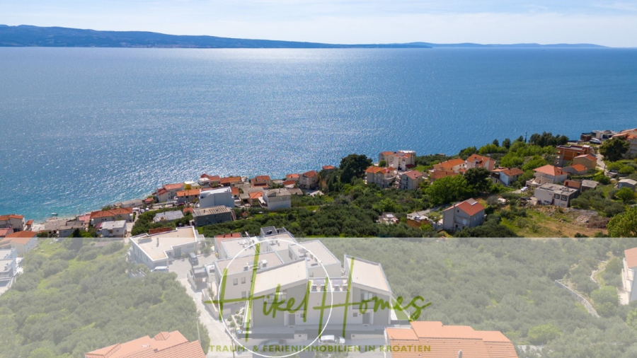 Direkt einziehen Meerblick Villa NATALIA mit Infinity Pool, 3 SZ, Sommerküche, … - Luftaufnahme eines Küstendorfes mit rot gedeckten Häusern, umgeben von Grünflächen an einem Hang mit Blick auf das ruhige, blaue Meer. In der Ferne sind Berge auf der anderen Seite des Wassers sichtbar. Das Bild ist unten mit einem Logo und Text überlagert. (43864)