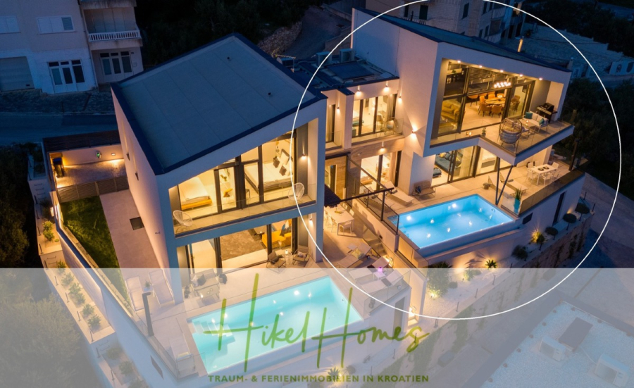 Direkt einziehen Meerblick Villa NATALIA mit Infinity Pool, 3 SZ, Sommerküche, … - Eine moderne, mehrstöckige Villa in Kroatien in der Abenddämmerung mit großen Fenstern, zwei beleuchteten Swimmingpools, Terrassen und Sitzgelegenheiten im Freien, von oben betrachtet. Der Text „Hikel Homes“ und „Traum- & Ferienimmobilien in Kroatien“ ist eingeblendet. (43873)