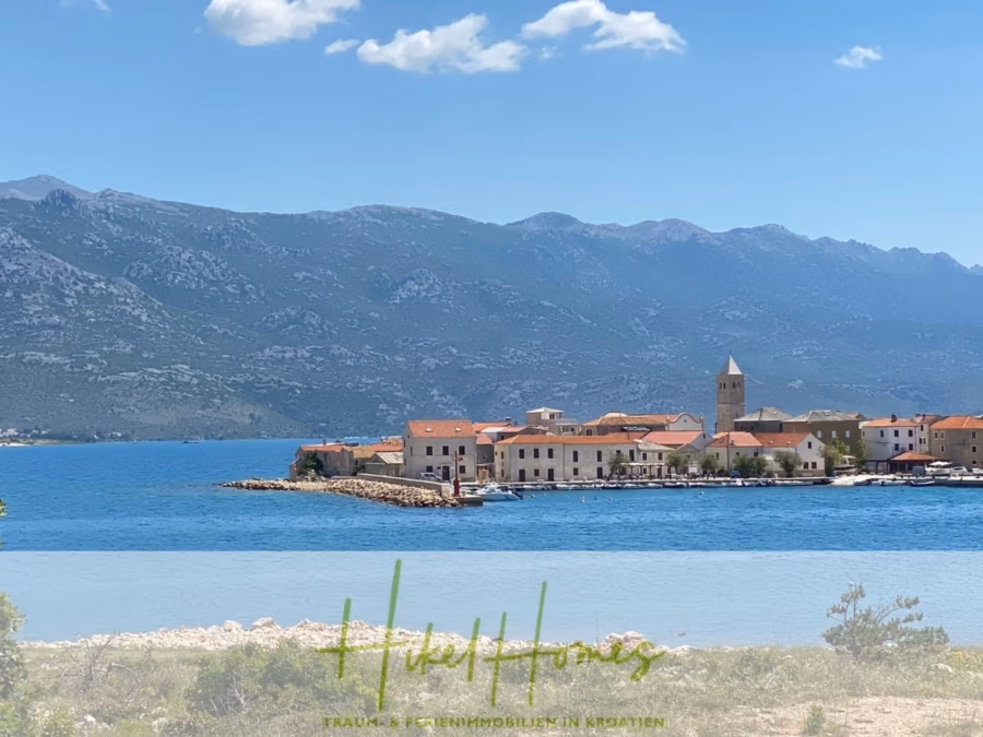 Nahe Zadar – Luxus-Villa in 2. Reihe zum Meer mit 4 SZ, Pool, Sauna, Fitness & Jacuzzi - Malerische Aussicht auf eine Küstenstadt mit rot gedeckten Gebäuden, eingebettet in das blaue Meer. Im Hintergrund erheben sich schroffe Berge unter einem klaren blauen Himmel. Ein kleiner Kirchturm ragt markant zwischen den Gebäuden hervor. Im Vordergrund ist Grün zu sehen. (42405)