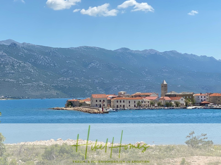 Nahe Zadar – Luxus-Villa in 2. Reihe zum Meer mit 4 SZ, Pool, Sauna, Fitness & Jacuzzi - Ein Küstendorf mit roten Dächern und einem Kirchturm liegt am blauen Wasser, umgeben von hohen Bergen unter einem klaren Himmel. Der Text "HuelHomes" ist am unteren Rand des Bildes zu sehen. (45976)