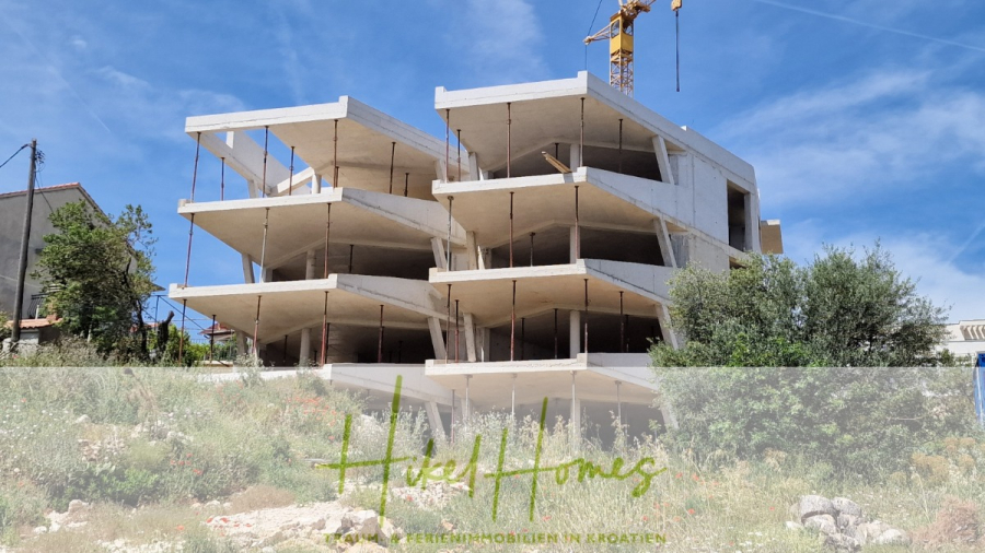 Meerblick Penthouse mit außergewöhnlicher Architektur 104m² mit 2 SZ, Terrasse und Südausrichtung - Ein modernes, unfertiges Betongebäude im Bau, mit mehreren Stockwerken und einem Kran darüber. Die Baustelle ist von Grünflächen und Wildpflanzen umgeben. Der Schriftzug "Heidi Homes" erscheint am unteren Bildrand. (46341)