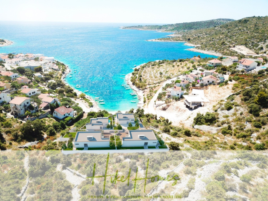 Villa Bettina – Luxus mit Blick auf Strand – Bucht – Meer / 3 Schlafzimmer / 3 Bäder - Luftaufnahme eines Küstendorfs mit Häusern, die eine türkisfarbene Bucht überblicken, umgeben von grünen Hügeln und felsiger Landschaft unter einem klaren blauen Himmel. (46769)