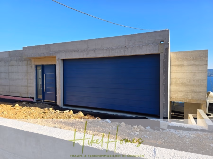 Schlüsselfertige – großzügige Meerblick Villa in ruhiger Lage, 4 SZ, 4 Bäder, Pool, … - Modernes Betonhaus mit Flachdach und großem blauen Garagentor auf einem unbebauten Grundstück. Das Haus verfügt über eine kleine angrenzende blaue Tür und steht unter einem strahlend blauen Himmel. Unten ist das Hikel Homes-Logo sichtbar. (42489)