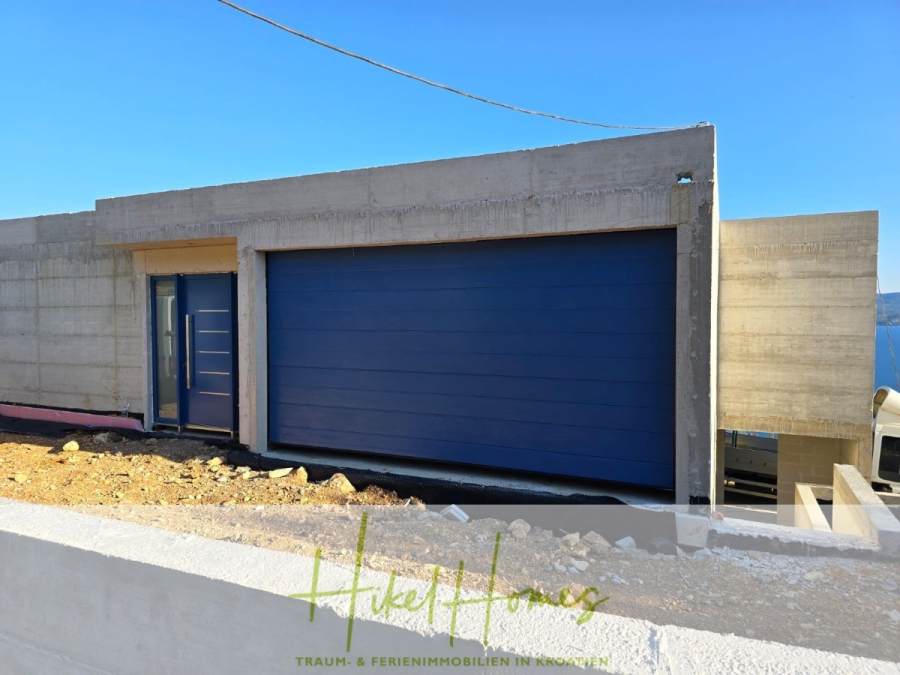 Schlüsselfertige – großzügige Meerblick Villa in ruhiger Lage, 4 SZ, 4 Bäder, Pool, … - Modernes Betonhaus mit einem großen blauen Garagentor und einer passenden blauen Eingangstür auf einem schrägen, unfertigen Grundstück unter einem klaren blauen Himmel. Der Schriftzug "Heike Homes" erscheint am unteren Rand mit deutscher Schrift. (45180)