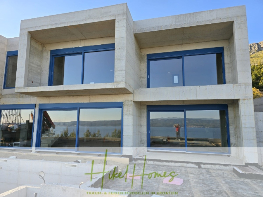 Schlüsselfertige – großzügige Meerblick Villa in ruhiger Lage, 4 SZ, 4 Bäder, Pool, … - Ein modernes, unfertiges Betonhaus mit großen, blau gerahmten Fenstern und Balkonen blickt auf das Meer, mit Bäumen und einem klaren Himmel im Hintergrund. Das Logo "Hikel Homes" befindet sich am unteren Rand des Bildes. (45176)