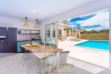 Moderne offene Küche und Essbereich mit Glasschiebetüren, die zu einer sonnigen Terrasse mit Pool, Liegestühlen und Sonnenschirmen führen. Der Raum ist hell, luftig und bietet einen Blick auf einen klaren blauen Himmel. - 4 Schlafzimmer / 32m² Pool / Sommerküche – Neubauhaus nur 8km von Omis entfernt