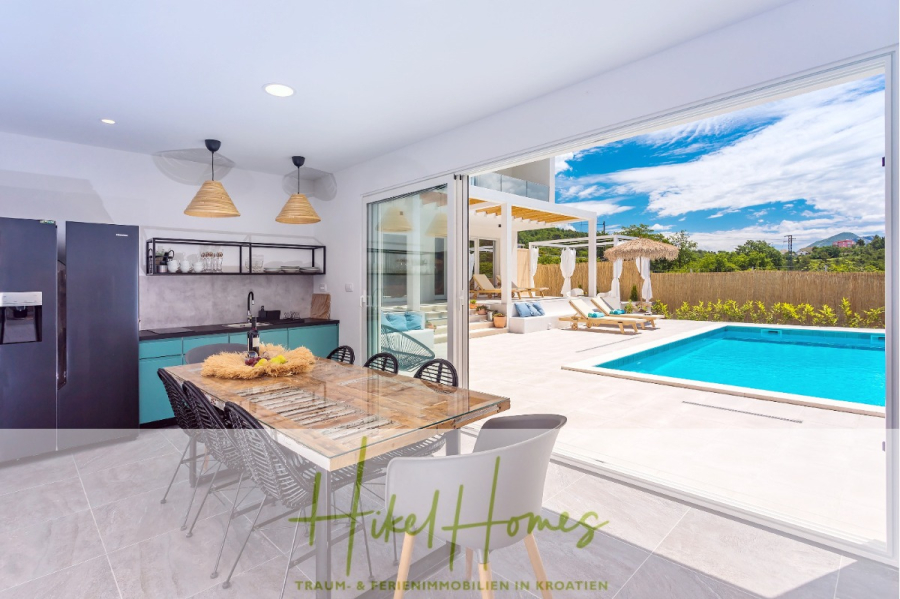 4 Schlafzimmer / 32m² Pool / Sommerküche – Neubauhaus nur 8km von Omis entfernt - Moderne offene Küche und Essbereich mit Glasschiebetüren, die zu einer sonnigen Terrasse mit Pool, Liegestühlen und Sonnenschirmen führen. Der Raum ist hell, luftig und bietet einen Blick auf einen klaren blauen Himmel. (44106)