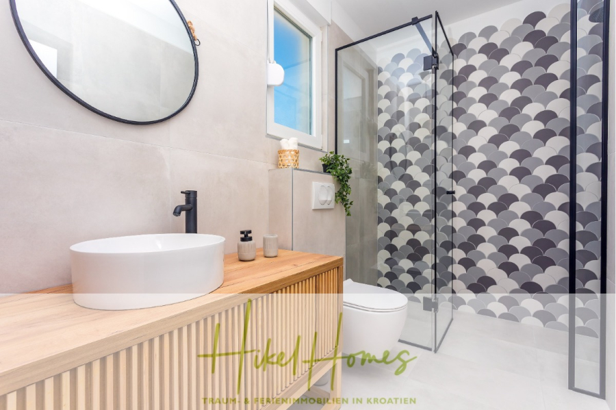 4 Schlafzimmer / 32m² Pool / Sommerküche – Neubauhaus nur 8km von Omis entfernt - Modernes Bad mit rundem Spiegel, Gefäßwaschbecken, schwarzem Wasserhahn, hölzernem Waschtisch und Glasdusche. Die Duschwand ist mit Fliesen mit Wellenmuster in Grautönen versehen. Ein Fenster lässt natürliches Licht herein. (44128)