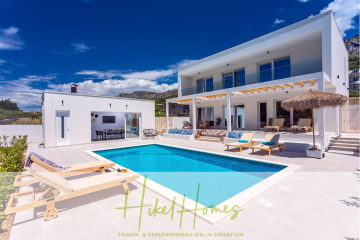 Moderne weiße Villa mit Swimmingpool, Liegestühlen und schattigem Sitzbereich im Freien vor der Kulisse von Hügeln unter einem strahlend blauen Himmel. Der Text "Hikel Homes" erscheint am unteren Rand. - 4 Schlafzimmer / 32m² Pool / Sommerküche – Neubauhaus nur 8km von Omis entfernt