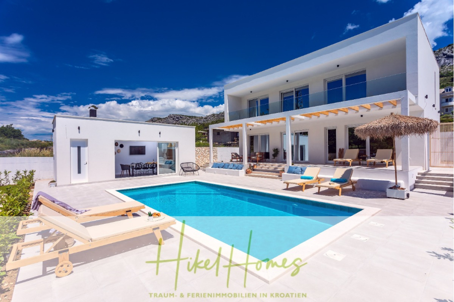 4 Schlafzimmer / 32m² Pool / Sommerküche – Neubauhaus nur 8km von Omis entfernt - Moderne weiße Villa mit Swimmingpool, Liegestühlen und schattigem Sitzbereich im Freien vor der Kulisse von Hügeln unter einem strahlend blauen Himmel. Der Text "Hikel Homes" erscheint am unteren Rand. (44103)