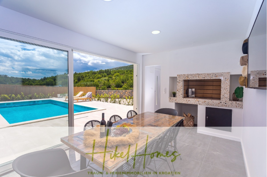 4 Schlafzimmer / 32m² Pool / Sommerküche – Neubauhaus nur 8km von Omis entfernt - Der moderne Essbereich mit Holztisch und schwarzen Stühlen bietet durch große Glastüren einen Blick auf den privaten Außenpool und die grüne Landschaft; der Küchenbereich verfügt über einen eingebauten Grill. (44108)