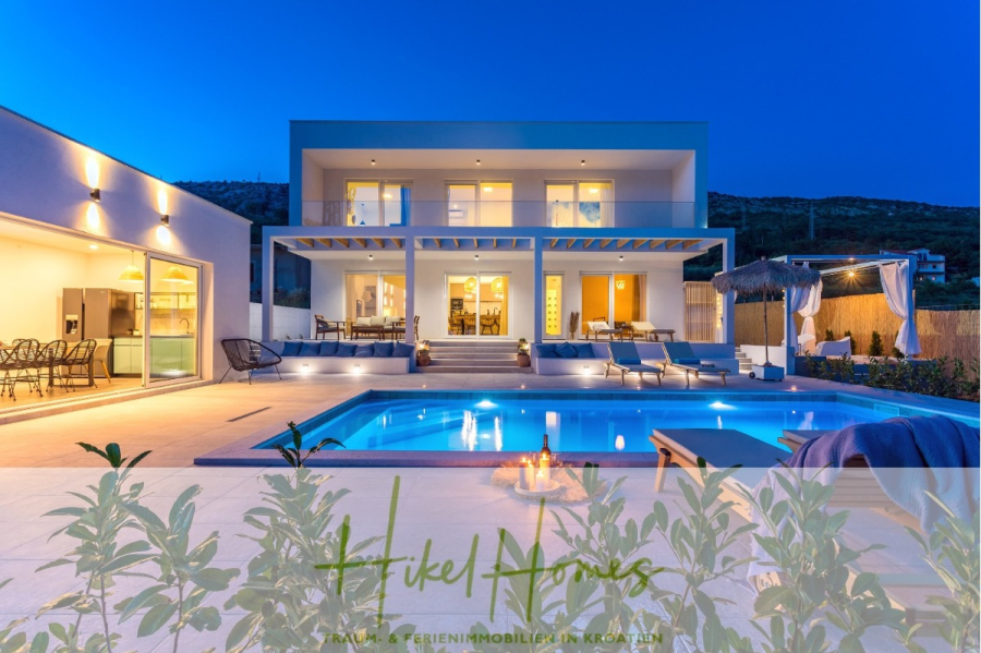 4 Schlafzimmer / 32m² Pool / Sommerküche – Neubauhaus nur 8km von Omis entfernt - Moderne zweistöckige Villa in der Abenddämmerung mit großen Fenstern, Sitzgelegenheiten im Freien, Liegestühlen am Pool, einem beleuchteten Swimmingpool und sanfter Außenbeleuchtung vor einer Hügelkulisse. Logo-Overlay am unteren Rand. (44136)