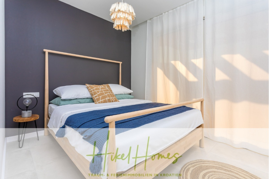 4 Schlafzimmer / 32m² Pool / Sommerküche – Neubauhaus nur 8km von Omis entfernt - Minimalistisches Schlafzimmer mit einem Bettgestell aus Holz, weißer und blauer Bettwäsche, einer kleinen Nachttischlampe auf einem runden Tisch, einer dunklen Akzentwand und transparenten Vorhängen, die das Sonnenlicht durchlassen. Eine geflochtene Pendelleuchte hängt über dem Bett. (44116)