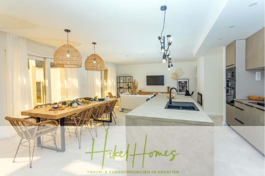 4 Schlafzimmer / 32m² Pool / Sommerküche – Neubauhaus nur 8km von Omis entfernt - Heller, moderner, offener Küchen- und Essbereich mit langem Holztisch, Korbstühlen, Pendelleuchten und einem gemütlichen Wohnbereich mit Sofa und Fernseher. Textüberlagerung: "Hike Homes" und "Traum- & Ferienimmobilien in Kroatien". (44111)