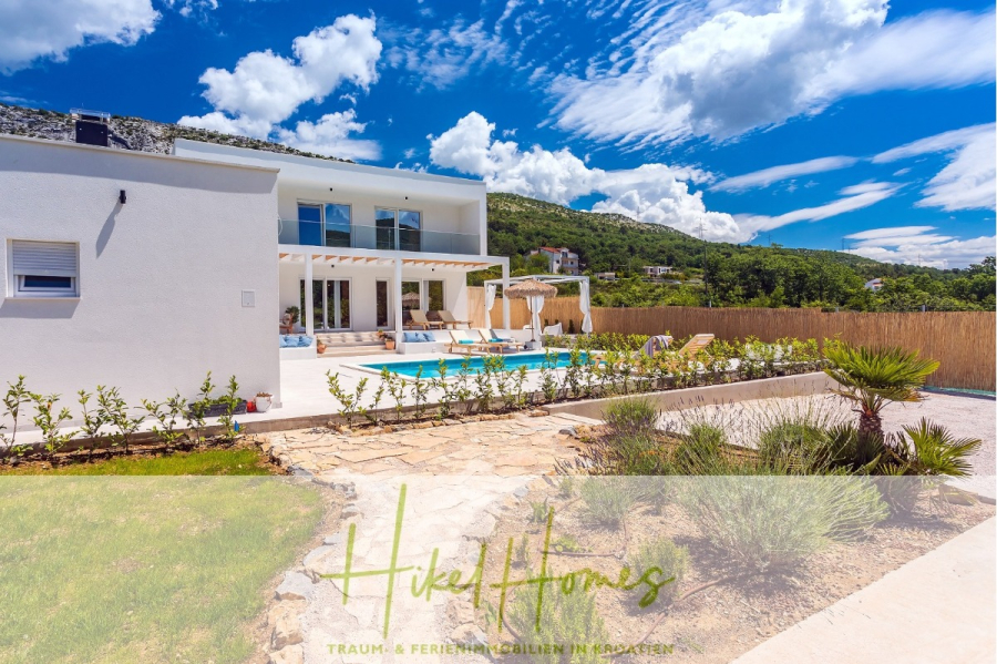 4 Schlafzimmer / 32m² Pool / Sommerküche – Neubauhaus nur 8km von Omis entfernt - Moderne weiße Villa mit Pool, Liegestühlen und einer schattigen Terrasse, umgeben von Grün und Hügeln unter einem blauen Himmel mit Wolken. Der Text "Hike Homes" und "TRAUM-4 FERIEN-VILLEN IN KROATIEN" erscheint am unteren Rand. (44131)