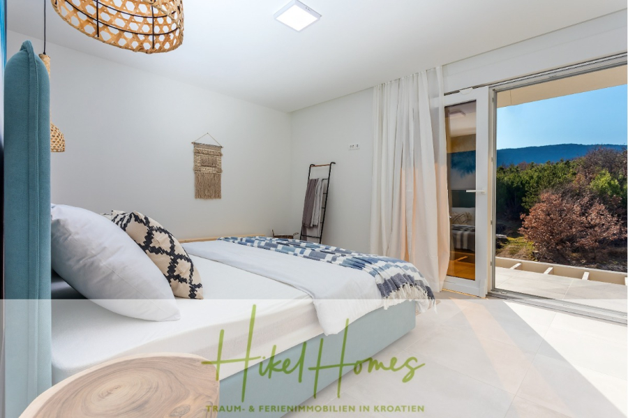 4 Schlafzimmer / 32m² Pool / Sommerküche – Neubauhaus nur 8km von Omis entfernt - Ein modernes Schlafzimmer mit einem großen Bett, einem hellblauen Kopfteil, gewebter Wandkunst und Glastüren, die zu einem Balkon mit malerischem Waldblick führen. Das Logo "Hikel Homes" ist am unteren Rand eingeblendet. (44123)