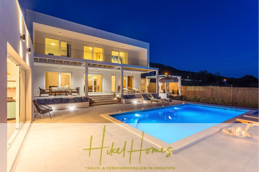 4 Schlafzimmer / 32m² Pool / Sommerküche – Neubauhaus nur 8km von Omis entfernt - Moderne zweistöckige Villa in der Abenddämmerung mit beleuchtetem Pool, Außenlounge, Essbereich und Balkonen. Umgeben von einem Sichtschutzzaun und Hügeln im Hintergrund. Der Text lautet "Hikel Homes - Traum- & Ferienimmobilien in Kroatien. (44134)