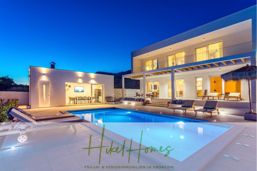 4 Schlafzimmer / 32m² Pool / Sommerküche – Neubauhaus nur 8km von Omis entfernt - Moderne Villa mit beleuchtetem Pool und geräumigem Patio in der Abenddämmerung, mit Sitzgelegenheiten im Freien, Essbereich und großen Glastüren. Das Haus ist zweistöckig, mit warmer Beleuchtung und modernem Design. Das Logo "Hikel Homes" ist auf dem Bild überlagert. (44133)