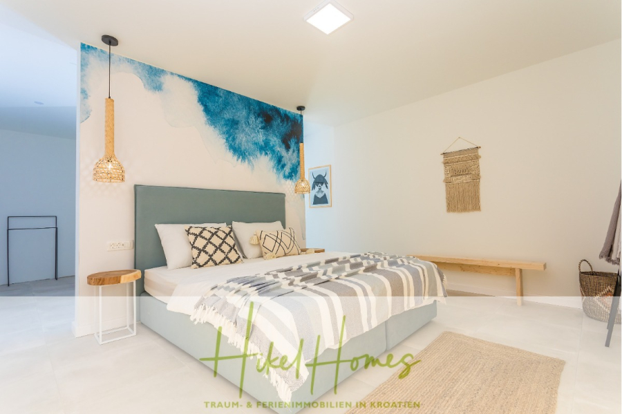 4 Schlafzimmer / 32m² Pool / Sommerküche – Neubauhaus nur 8km von Omis entfernt - Ein modernes Schlafzimmer mit einem großen Bett, blauen Akzenten an der Wand, hängenden Pendelleuchten, einer Holzbank und minimalem Dekor. Der Raum ist hell, luftig und modern. Der Schriftzug "Hikel Homes" befindet sich am unteren Rand des Bildes. (44124)
