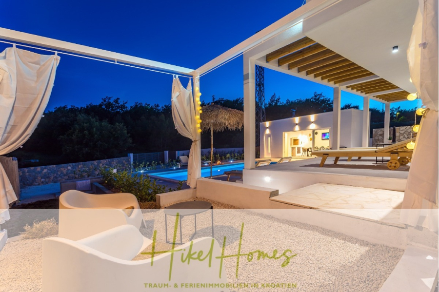 4 Schlafzimmer / 32m² Pool / Sommerküche – Neubauhaus nur 8km von Omis entfernt - Eine moderne Außenterrasse in der Abenddämmerung mit weißen Sitzgelegenheiten, einer Pergola mit Vorhängen, einem Pool und einem beleuchteten Garten. "Hikel Homes" und "Traum- & Ferienimmobilien Kroatien" stehen am unteren Rand des Bildes. (44135)