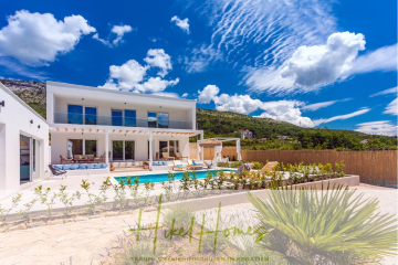 Moderne weiße Villa mit großen Fenstern, Außenterrasse, Pool und Sonnenliegen vor einer Kulisse grüner Hügel unter einem strahlend blauen Himmel mit vereinzelten Wolken. Zierpflanzen säumen den Vordergrund. - 4 Schlafzimmer / 32m² Pool / Sommerküche – Neubauhaus nur 8km von Omis entfernt