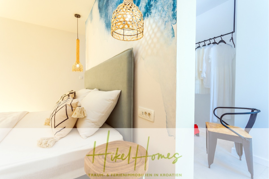 4 Schlafzimmer / 32m² Pool / Sommerküche – Neubauhaus nur 8km von Omis entfernt - Helles, modernes Schlafzimmer mit einem hellgrünen Kopfteil, gemusterten Kissen, Rattan-Pendelleuchten und einer blauen Akzentwand; angrenzender Ankleidebereich mit schwarzem Stuhl und weißer Hängegarderobe. "Hiky Homes"-Logo am unteren Rand. (44126)